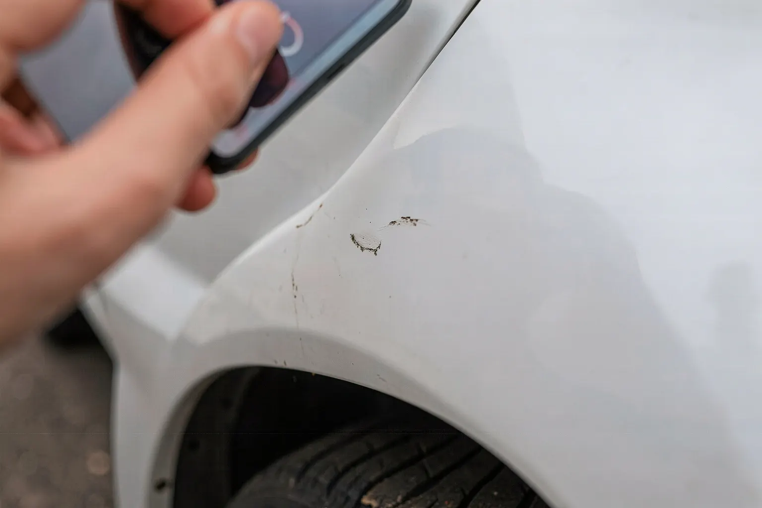 Gros plan sur un bas de porte et un passage de roue d'une voiture blanche portant une petite éraflure, smartphone tenu à la main pour cadrer la marque