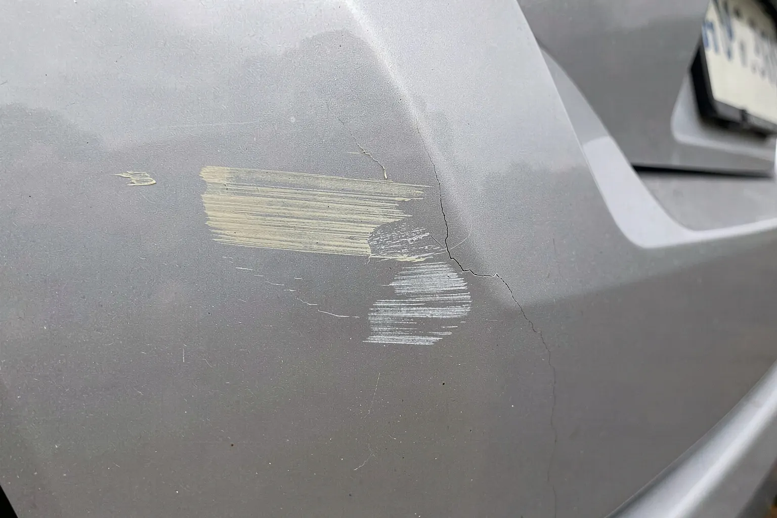 Gros plan sur un pare-chocs arrière gris métallisé portant une trace de peinture claire et une fissure fine, sous un ciel couvert