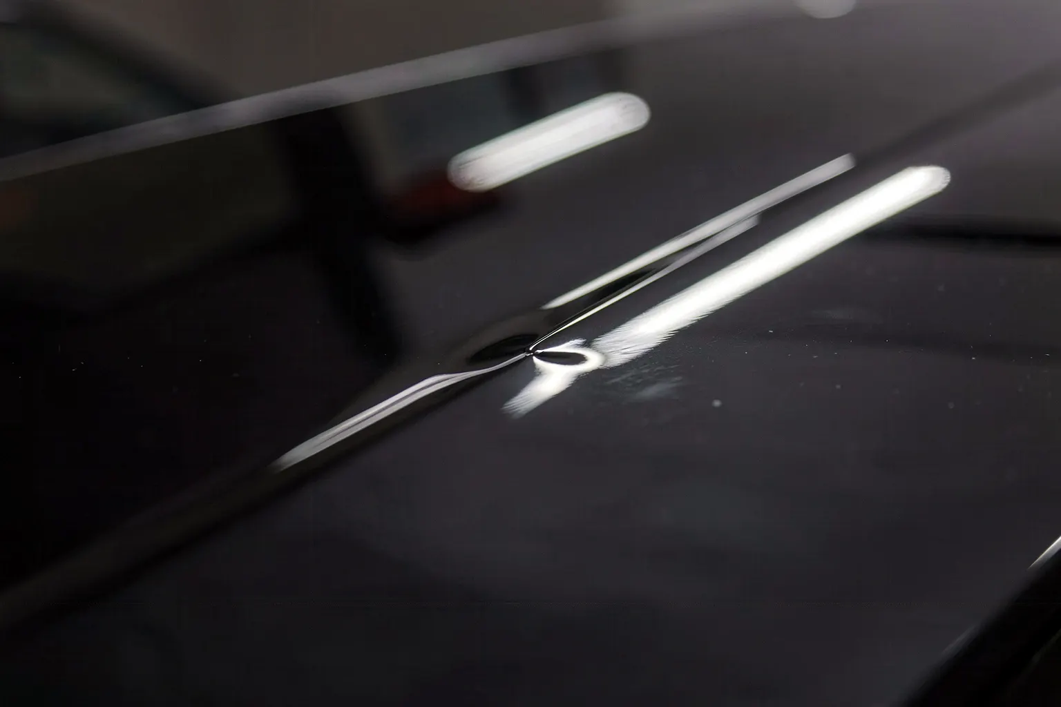 Gros plan sur la peinture noire brillante d'un capot où un petit creux plie le reflet allongé d'une bande de fenêtre